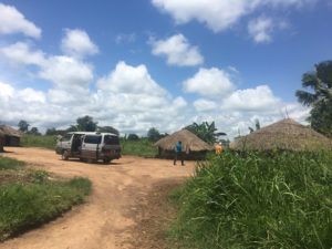 Old minivan with side door open at the entrance of a dirt road into a village with thatched roof huts and two tall figures walking in the distance